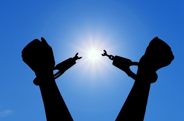 Silhouette of hands breaking free from handcuffs against a bright sun in a clear blue sky.