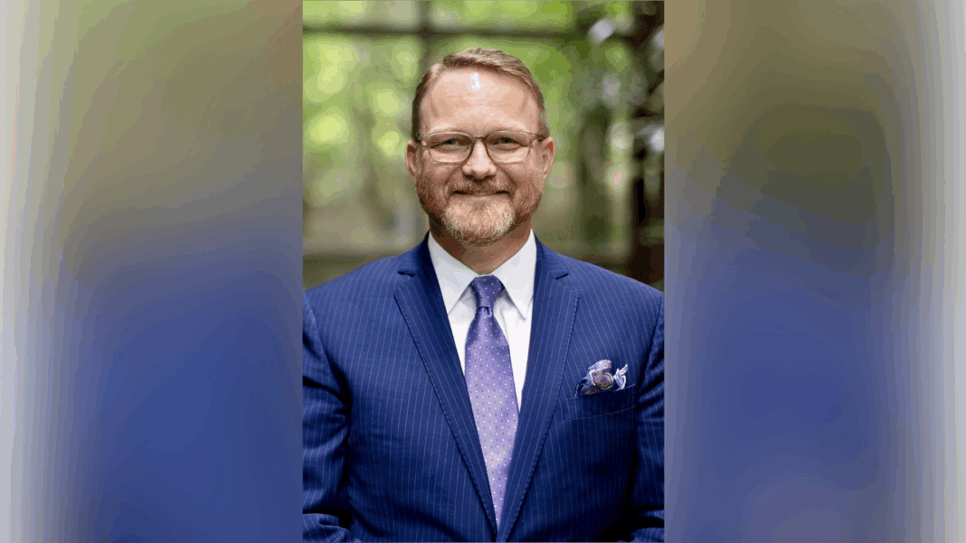 A man wearing glasses, a blue suit, and a purple tie stands indoors, smiling at the camera.