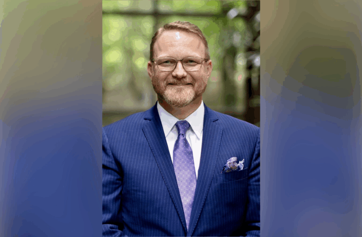 A man wearing glasses, a blue suit, and a purple tie stands indoors, smiling at the camera.
