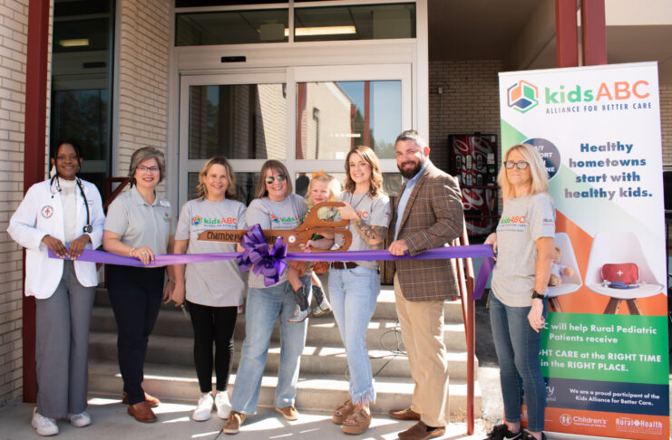 Seven people stand behind a purple ribbon at an outdoor event in front of a building, with a kids ABC banner displayed.