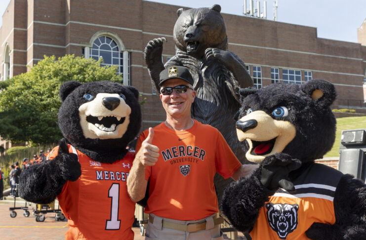 A person in a Mercer University shirt poses with two bear mascots in front of a bear statue on campus.