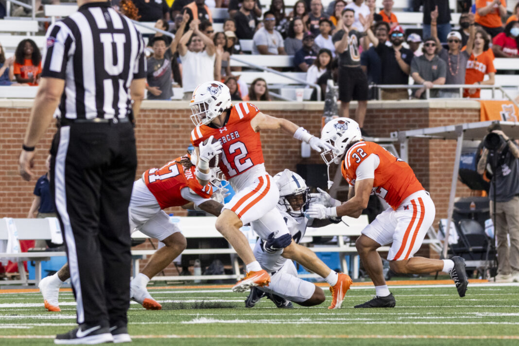 2025FootballMercerSamfordJessicaGratigny454 A football player in an orange jersey runs with the ball as two teammates block and a referee stands nearby.