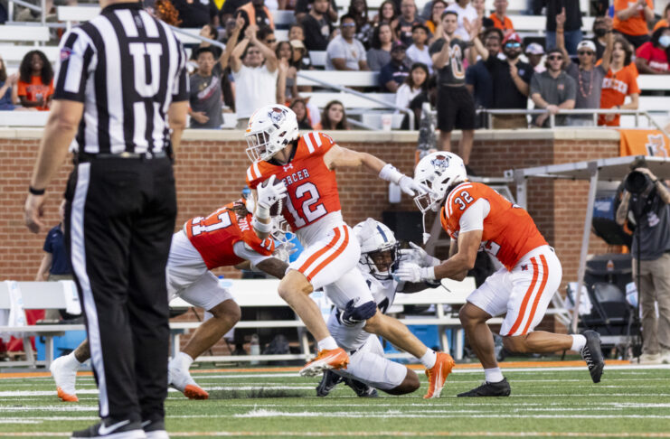 A football player in an orange jersey runs with the ball as two teammates block and a referee stands nearby.