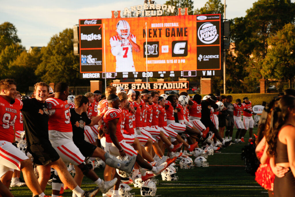 Mercer University football players link arms and kick in unison on the field before a game at Five Star Stadium.