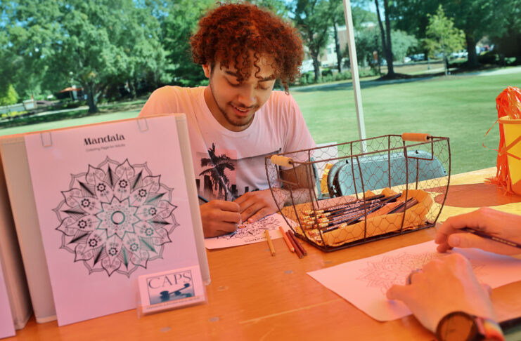 A man sits at an outdoor table coloring a mandala design with colored pencils beside a basket of art supplies.