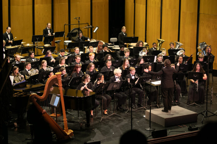 A conductor leads a large orchestra of musicians playing various instruments on a concert hall stage.