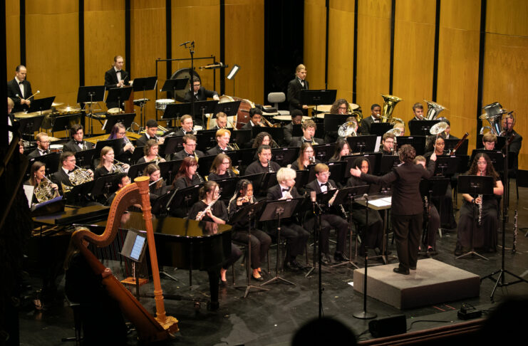 A conductor leads a large orchestra of musicians playing various instruments on a concert hall stage.