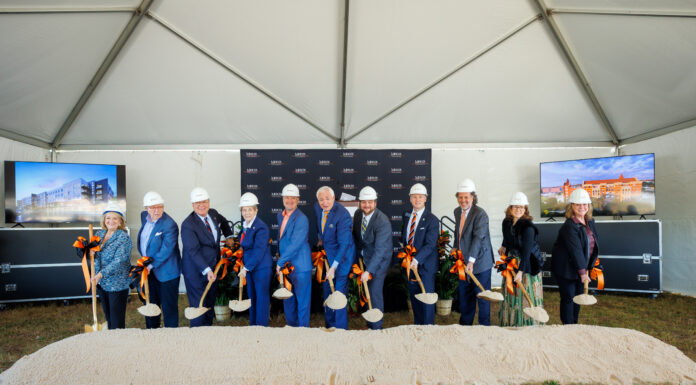 Mercer breaks ground on new medical school building in downtown Macon Eleven people in hard hats hold shovels at a groundbreaking ceremony under a tent with two display screens and a sand pile.