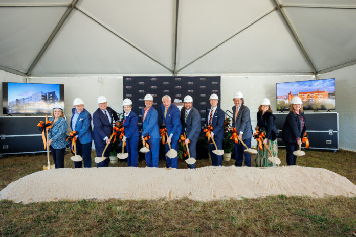 Eleven people in hard hats hold shovels at a groundbreaking ceremony under a tent with two display screens and a sand pile.