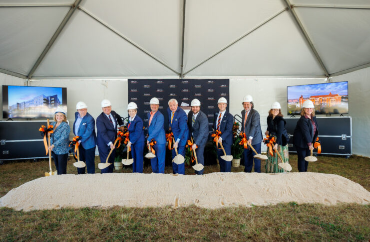 Eleven people in hard hats hold shovels at a groundbreaking ceremony under a tent with two display screens and a sand pile.
