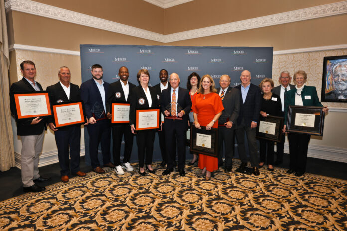 251114AlumniAwards_5883 A group of people stand indoors holding awards and framed certificates in front of a Mercer University backdrop.