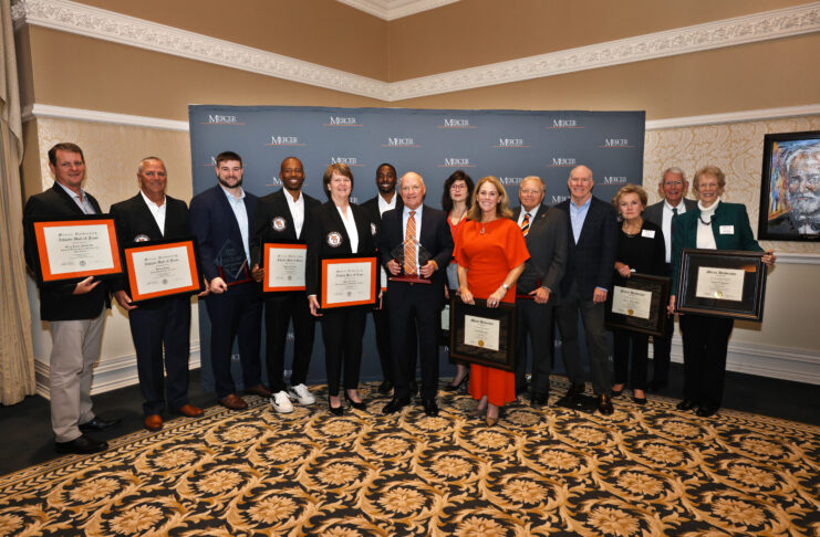 A group of people stand indoors holding awards and framed certificates in front of a Mercer University backdrop.