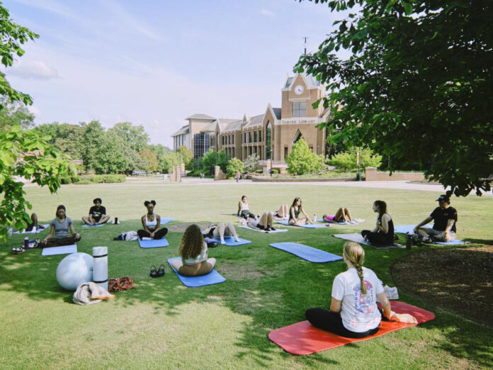 36B885F9-57DB-4DA0-ADF8-AE4D5A07EDAD A group of people practice yoga on mats in a grassy area near a large brick building on a sunny day.