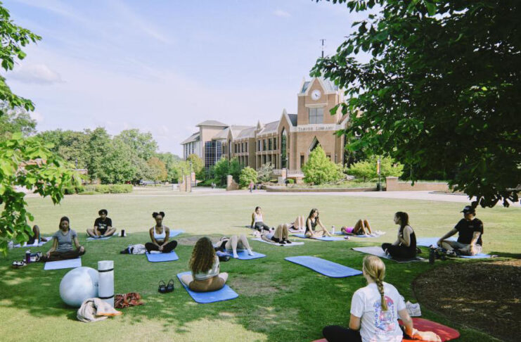 A group of people practice yoga on mats in a grassy area near a large brick building on a sunny day.