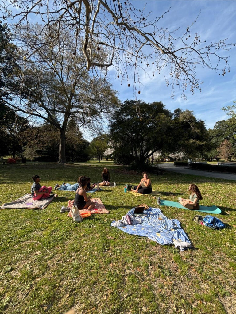 Six people sit on blankets and mats in a grassy park, spaced apart, with trees and blue sky in the background.