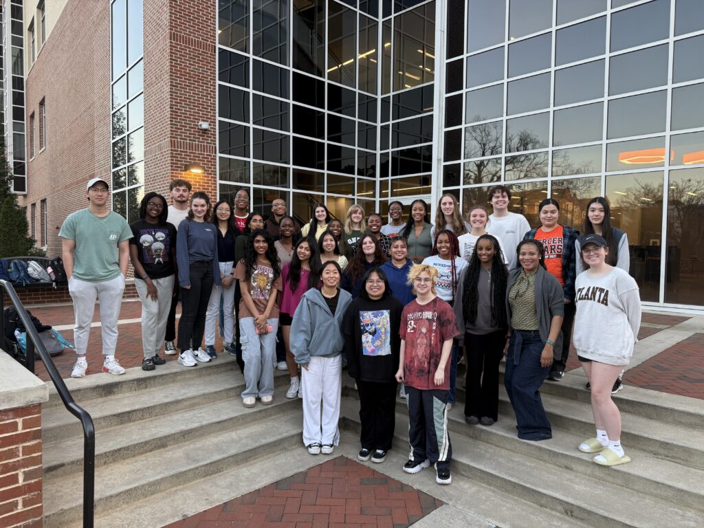 A group of students poses on outdoor steps in front of a modern building with large glass windows at dusk.