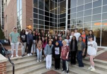 Biology professor recognized for student mentorship initiatives A group of students poses on outdoor steps in front of a modern building with large glass windows at dusk.