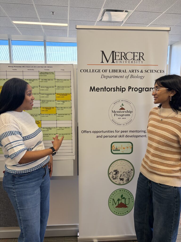 Two women stand by a mentorship program banner and a schedule, discussing information at Mercer University’s Department of Biology.