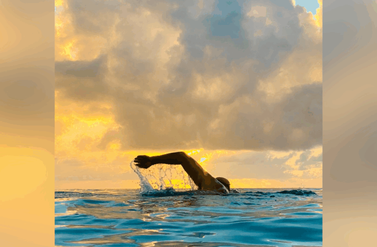 A swimmer performs a freestyle stroke in open water at sunset, with clouds and golden light in the sky.