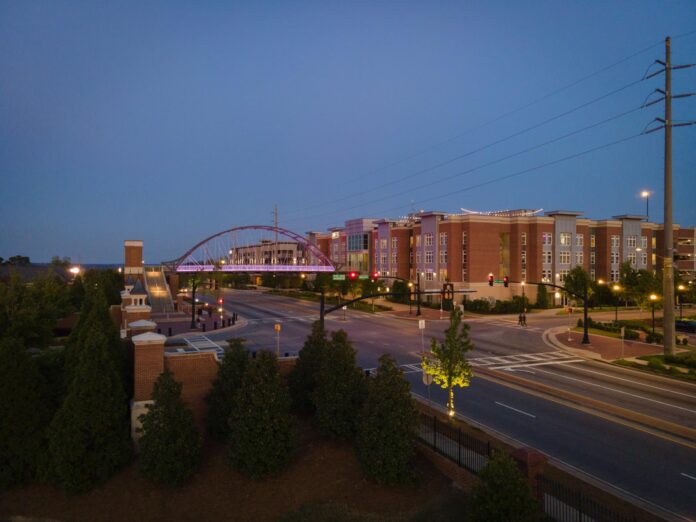 A street scene at dusk shows a pedestrian bridge, modern buildings, and trees, with streetlights and sparse traffic.