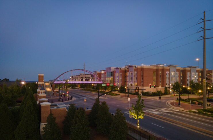 A street scene at dusk shows a pedestrian bridge, modern buildings, and trees, with streetlights and sparse traffic.