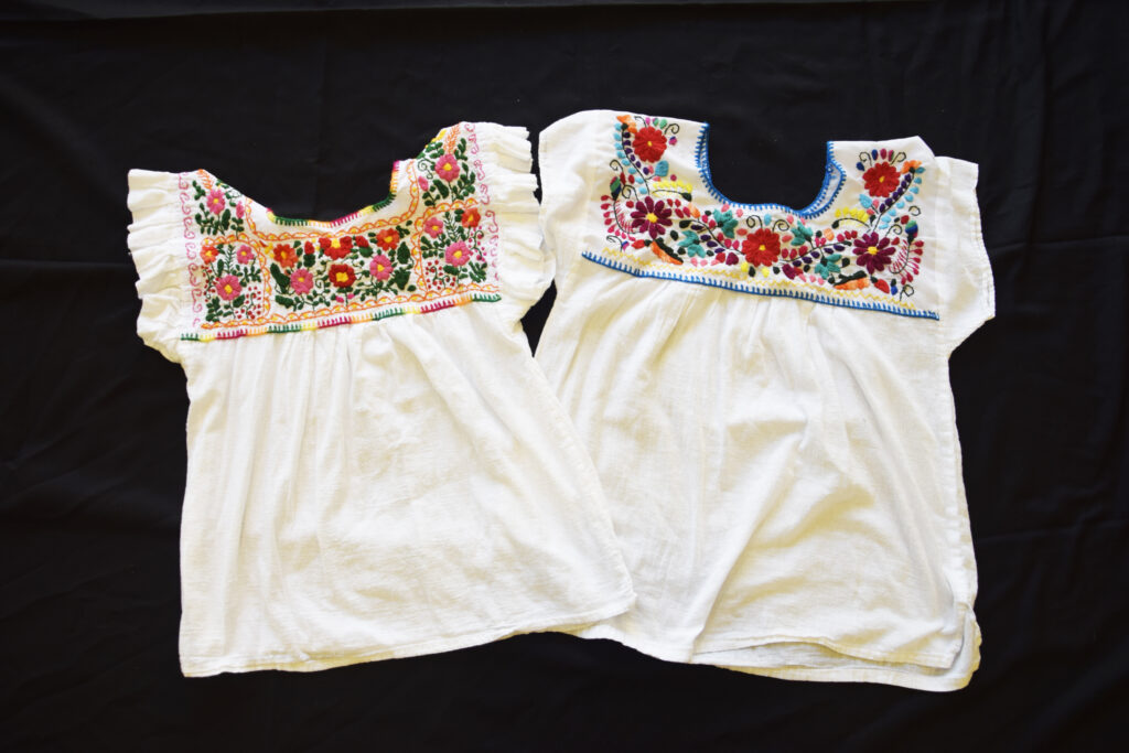 Two white, short-sleeved blouses with colorful floral embroidery on the yokes displayed on a black background.