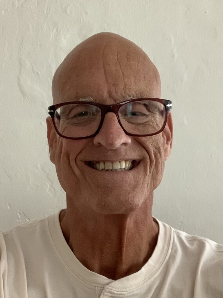 A smiling bald man wearing glasses and a white shirt stands in front of a plain, light-colored wall.