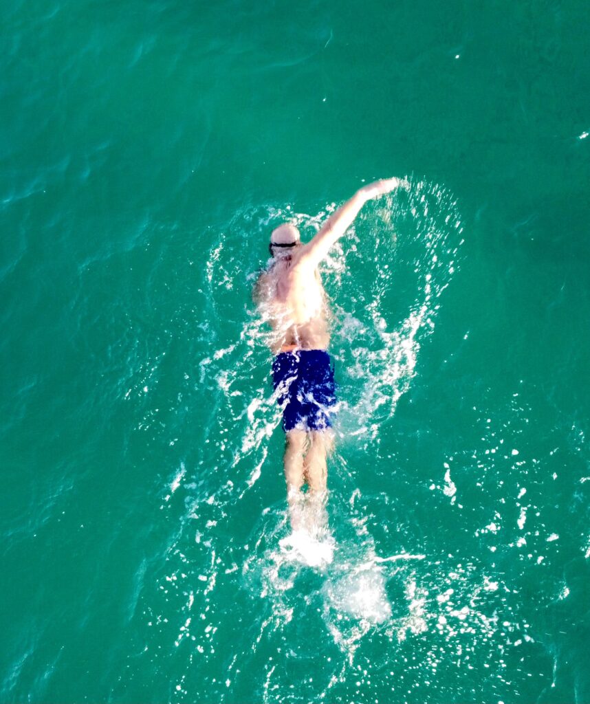 A person wearing swim goggles and blue shorts swims freestyle in clear turquoise water.