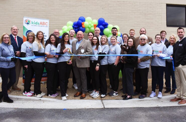 A group of people pose for a ribbon-cutting ceremony outside a building, with balloons and a kidsABC banner in the background.