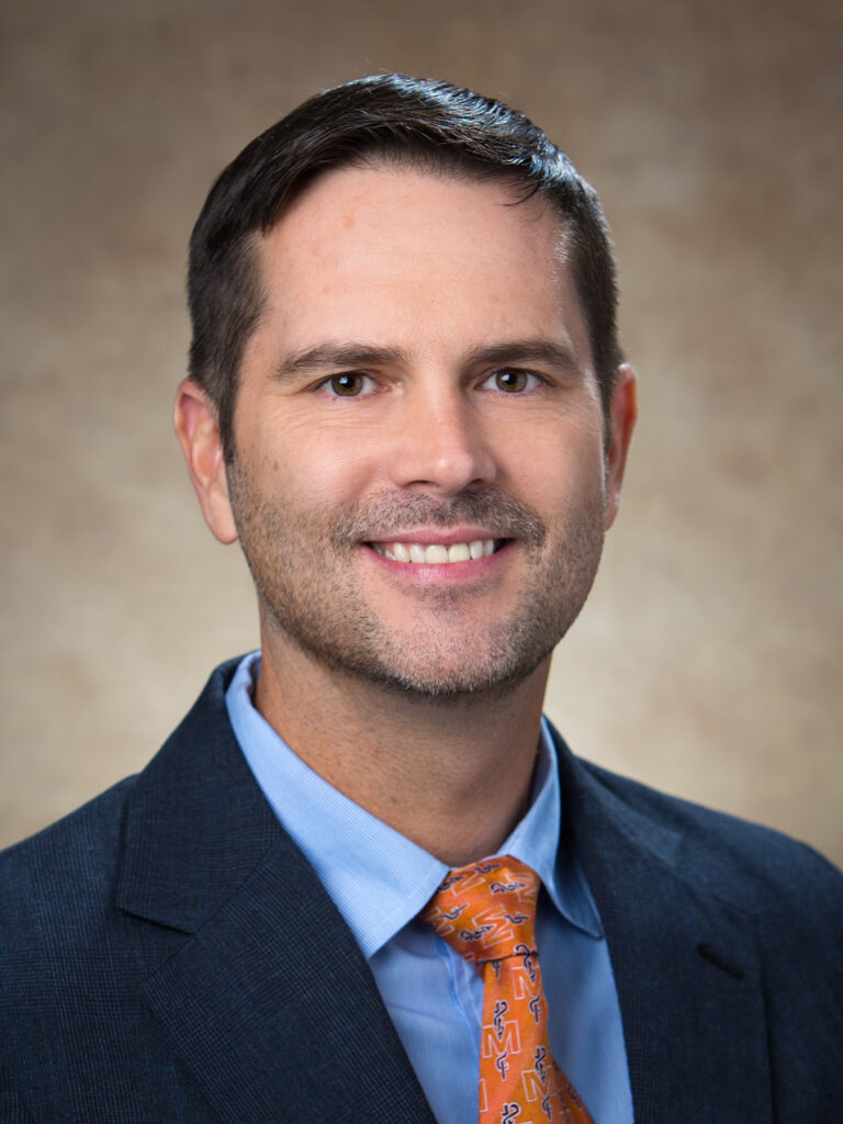 Dr. Joe Hayes in a suit and orange patterned tie smiling in a formal portrait with a brown background.