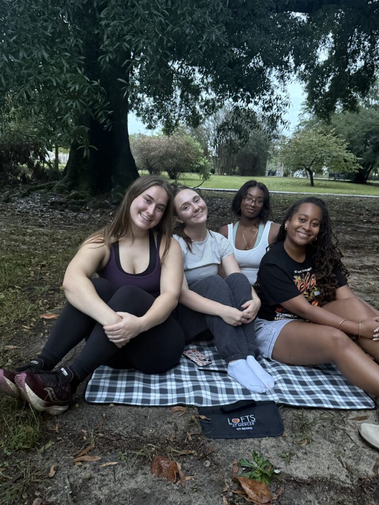 Four women sit on a plaid blanket outdoors in a park, smiling at the camera under trees on a cloudy day.