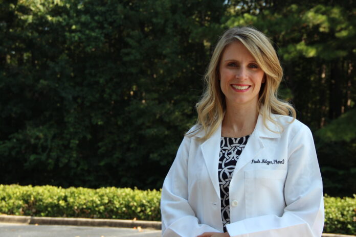 Woman in a white lab coat standing outdoors with trees and greenery in the background.