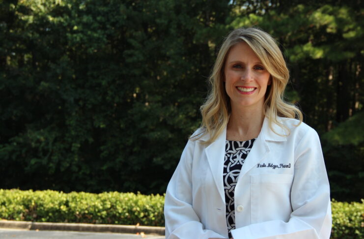 Woman in a white lab coat standing outdoors with trees and greenery in the background.