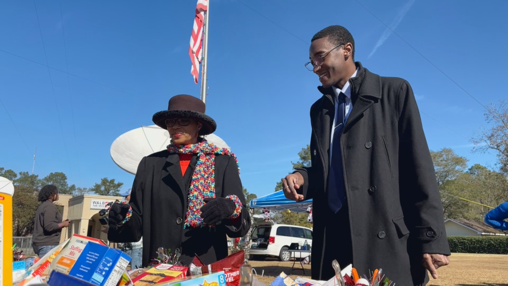Two people in coats stand at an outdoor table with snacks, with an American flag and satellite dishes in the background.