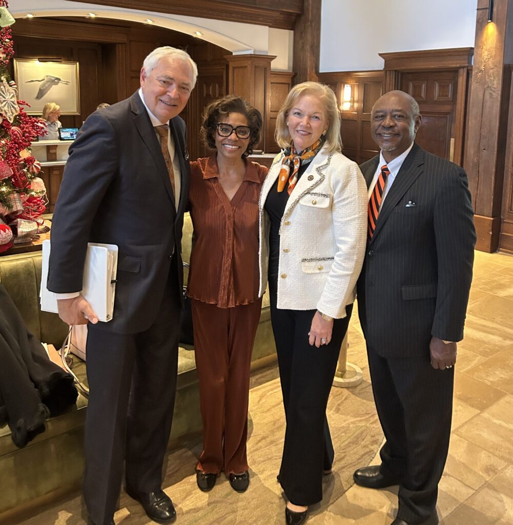 Four adults in business attire stand together and smile for a group photo in a wood-paneled, decorated lobby.