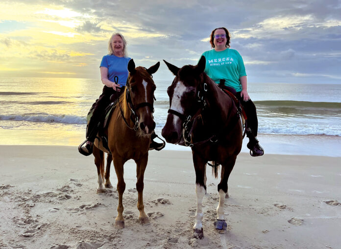 Two women riding horses on a sandy beach with the ocean and a cloudy sky in the background at sunset.
