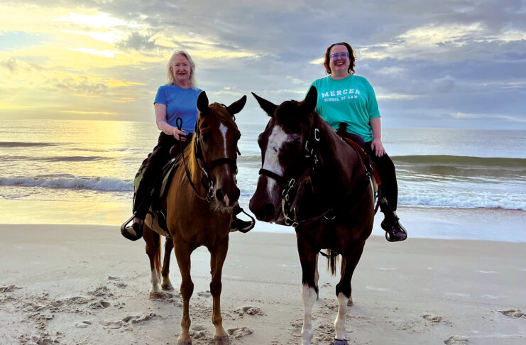 Two women riding horses on a sandy beach with the ocean and a cloudy sky in the background at sunset.