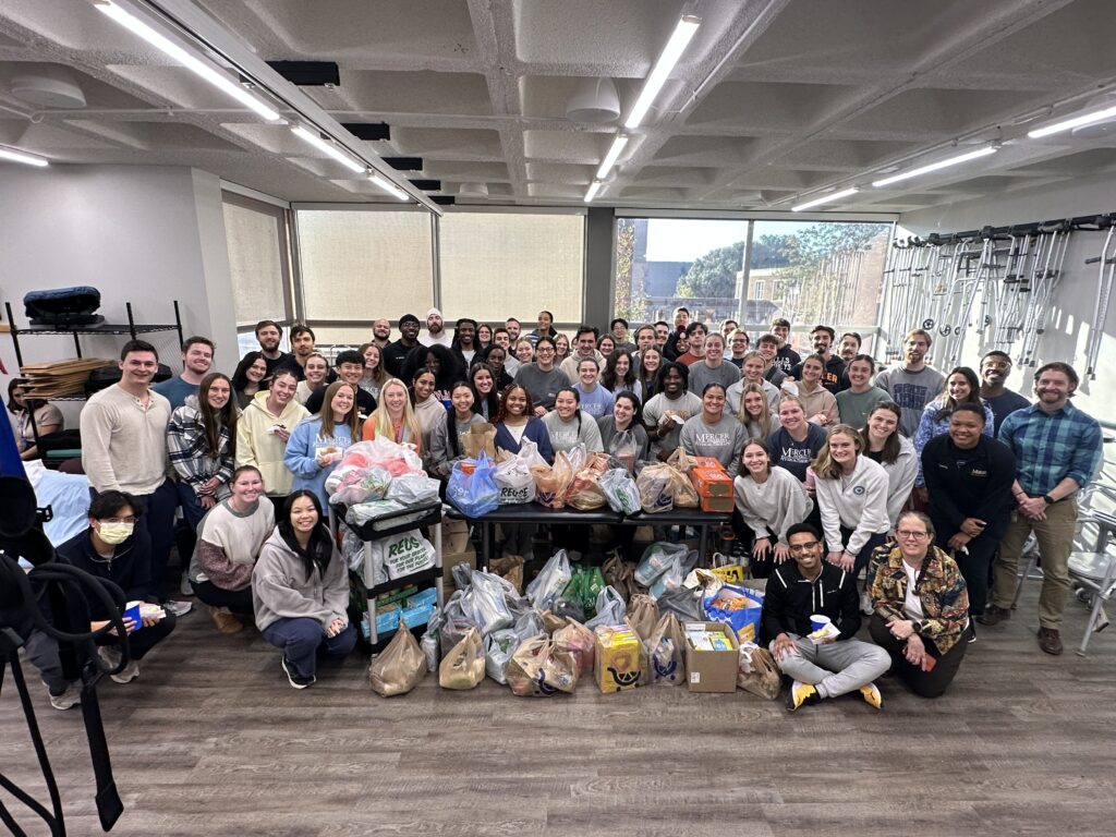A large group of people poses indoors around tables filled with assorted bags and boxes of donated goods.