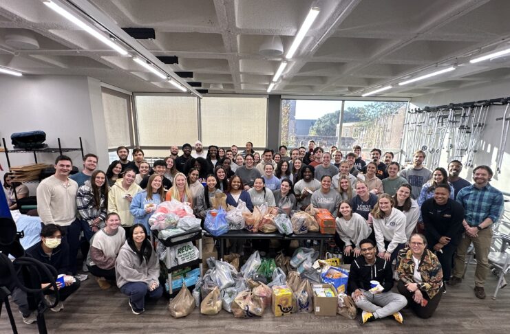 A large group of people poses indoors around tables filled with assorted bags and boxes of donated goods.