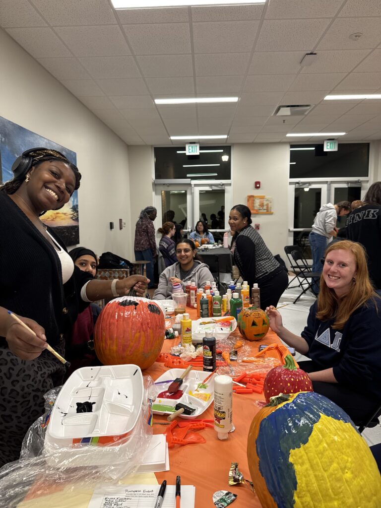 A group of people paint pumpkins at a table covered with art supplies in a well-lit indoor space.