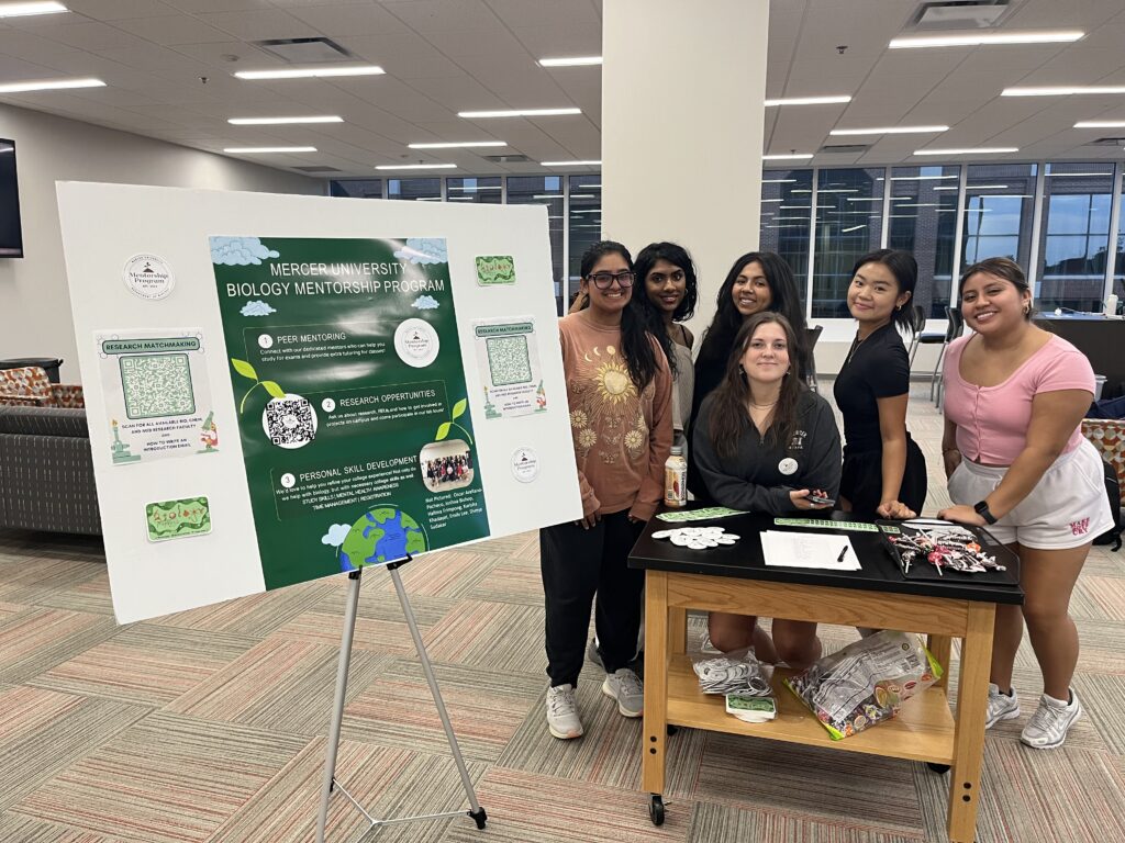 Six students stand by a poster display for a university biology mentorship program in a library or lounge area.