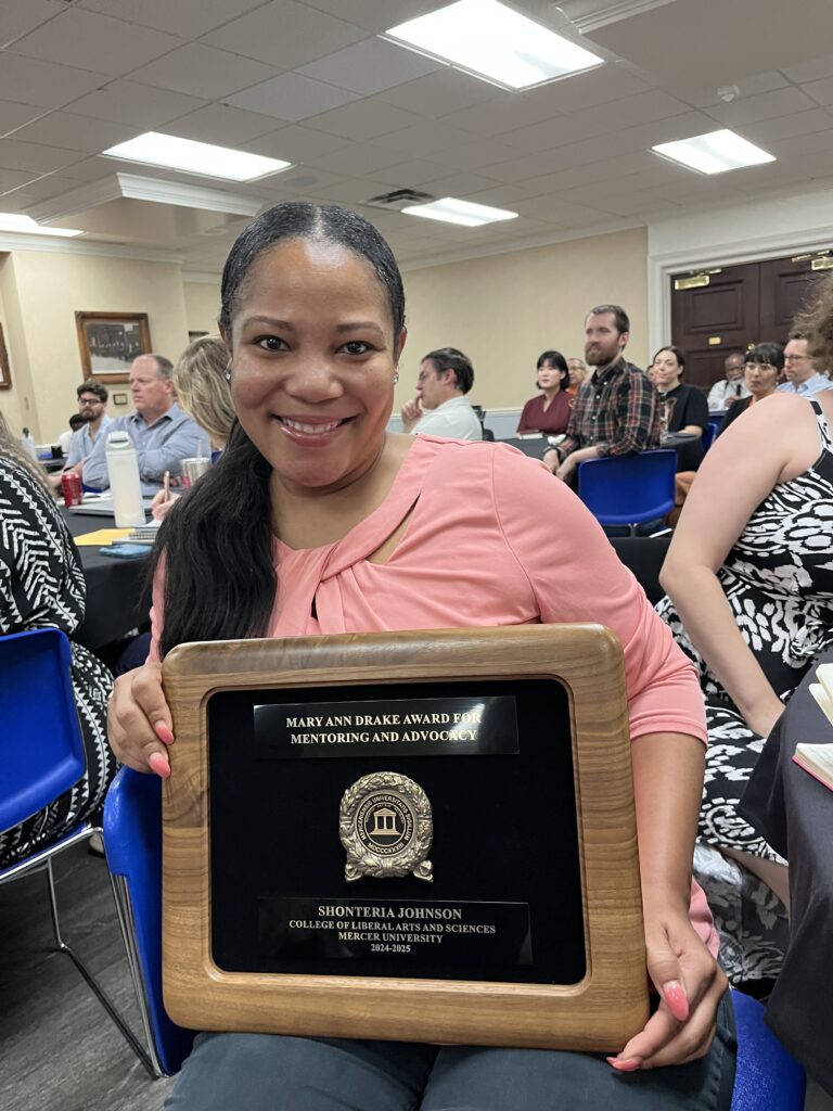 Woman smiling and holding a framed award plaque at an indoor event with people seated in the background.