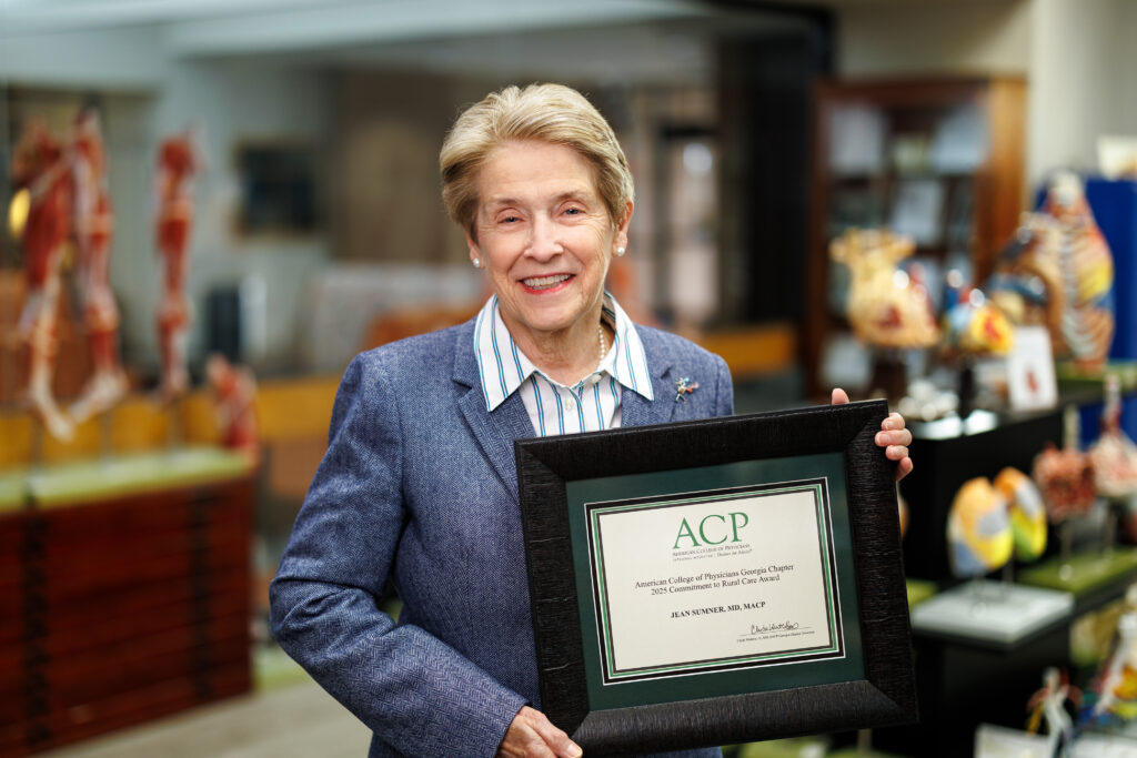 Dr. Jean Sumner holds a framed ACP certificate, standing in the MUSM medical library