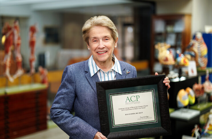 Dr. Jean Sumner holds a framed ACP certificate, standing in the MUSM medical library