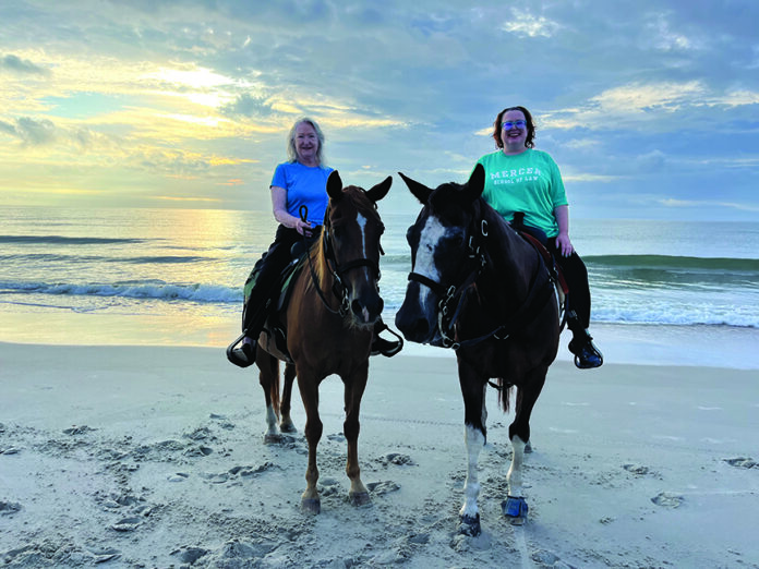 Two women riding horses on a sandy beach with the ocean and a cloudy sky in the background at sunset.