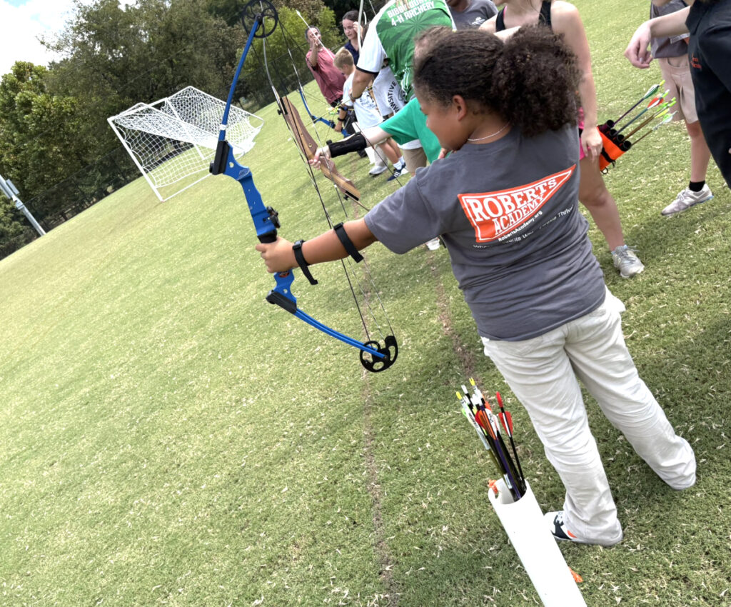 Children standing in a line outdoors practice archery, aiming bows at targets on a grassy field near soccer goals.