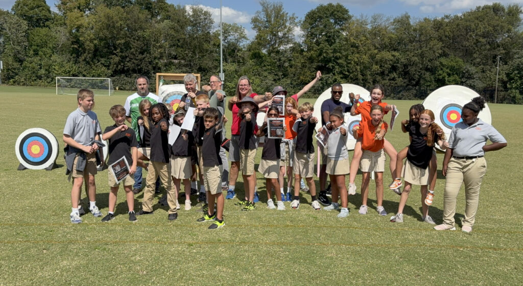 A group of students and adults pose outdoors on a field near archery targets under a sunny sky.