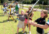 Taking aim at opportunity: A Mercer lawyer’s mission to empower youth through archery Children and adults practice archery outdoors on a grassy field, aiming bows at targets in the distance.
