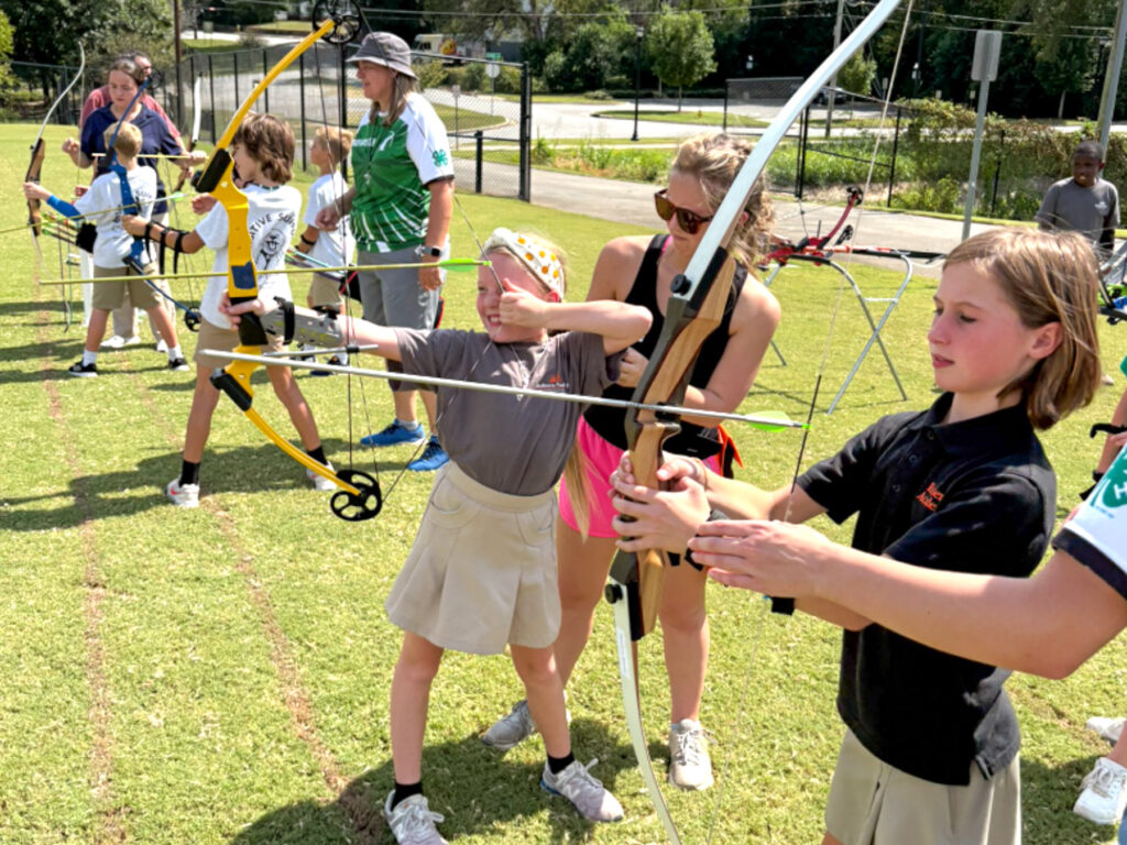Children and adults practice archery outdoors on a grassy field, aiming bows at targets in the distance.