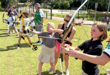 Taking aim at opportunity: A Mercer lawyer’s mission to empower youth through archery Children and adults practice archery outdoors on a grassy field, aiming bows at targets in the distance.